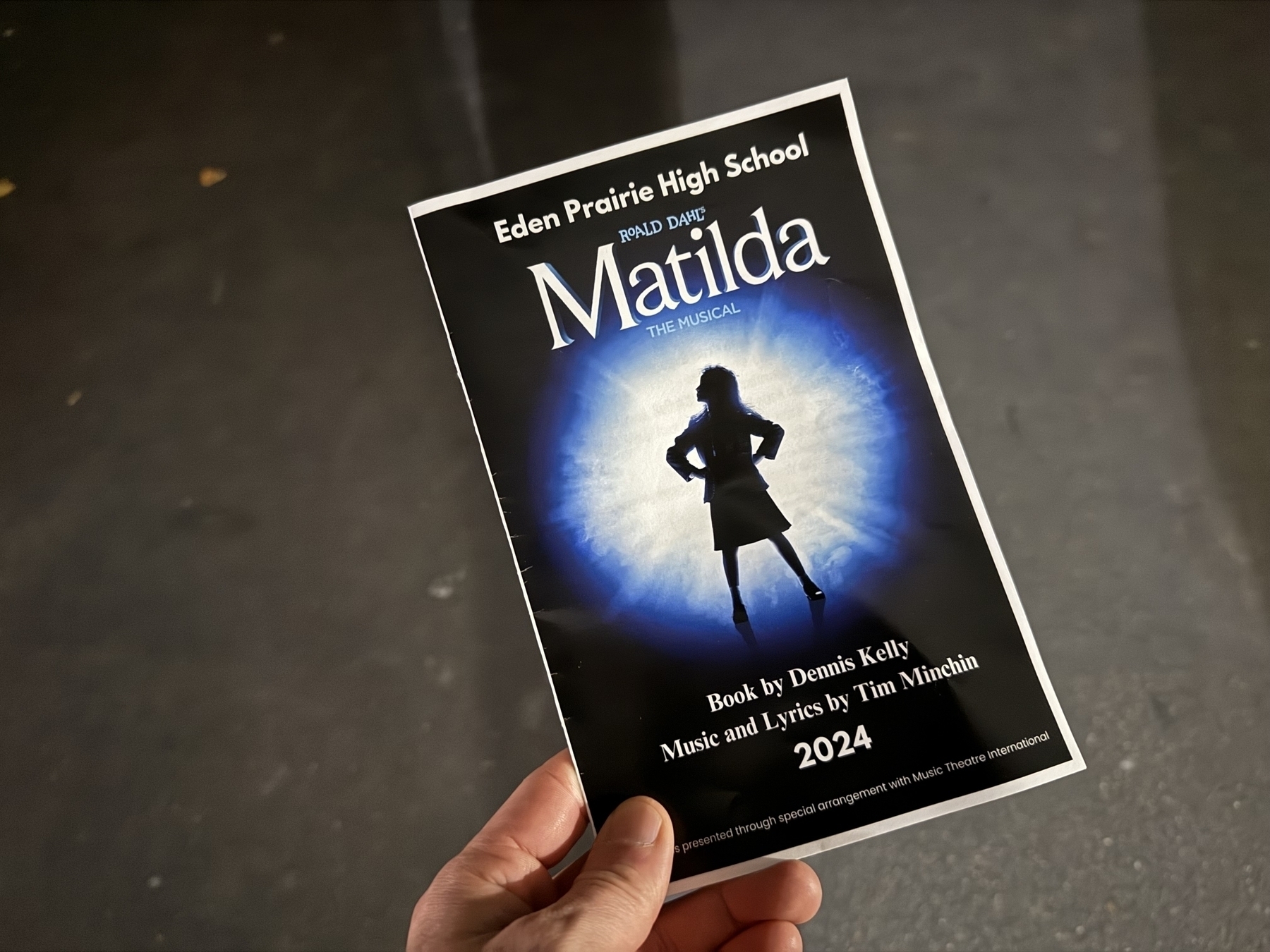 A hand holds a “Matilda the Musical” program against a dark floor. The cover features a silhouetted girl in front of a glowing blue background. Text: “Eden Prairie High School,” “2024,” “Book by Dennis Kelly, Music and Lyrics by Tim Minchin.