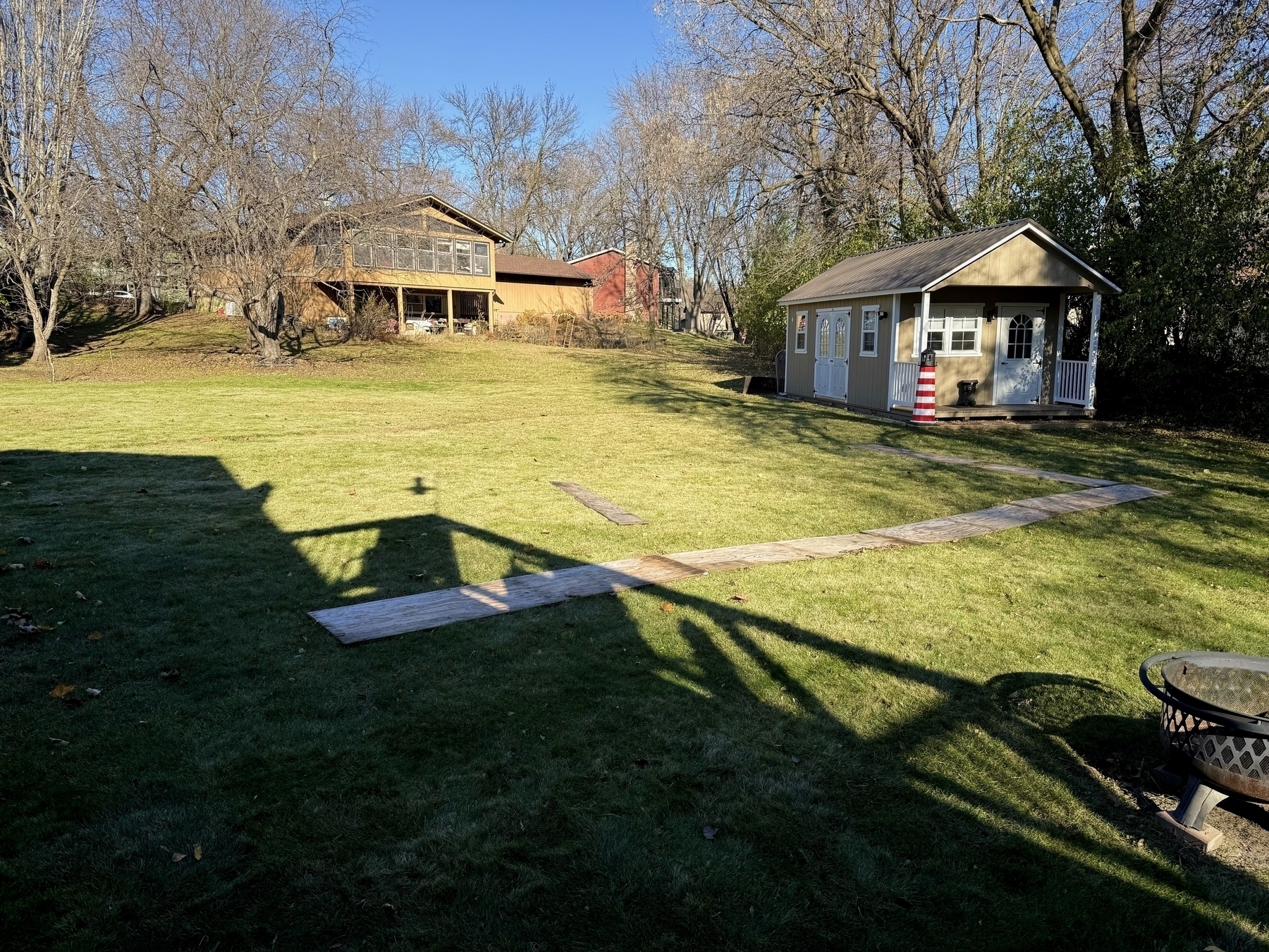 A small garden shed stands on a manicured lawn, casting long shadows on a sunny day, flanked by leafy trees with a wooden house visible in the background.